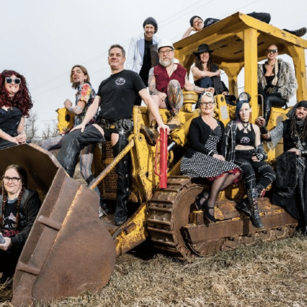 A diverse group of twelve people poses on and around a yellow bulldozer in an outdoor setting reminiscent of a Colorado Burning Man event. They wear eclectic outfits, some with tattoos and colorful hair, against a backdrop of wood piles and utility poles.